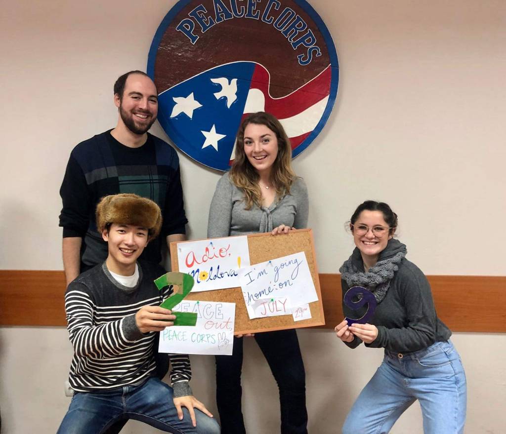 Four individuals (two male and two female) smile at the camera. In the middle of the group is a sign that reads "Adio Moldova" and "I'm going home on July 29."